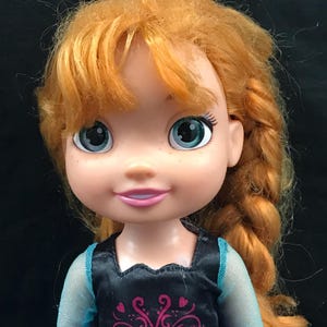 May include: A doll with orange hair styled in a side braid. The doll has blue eyes and is wearing a black top with a pink and purple design and a blue dress. The doll's skin is fair with freckles.