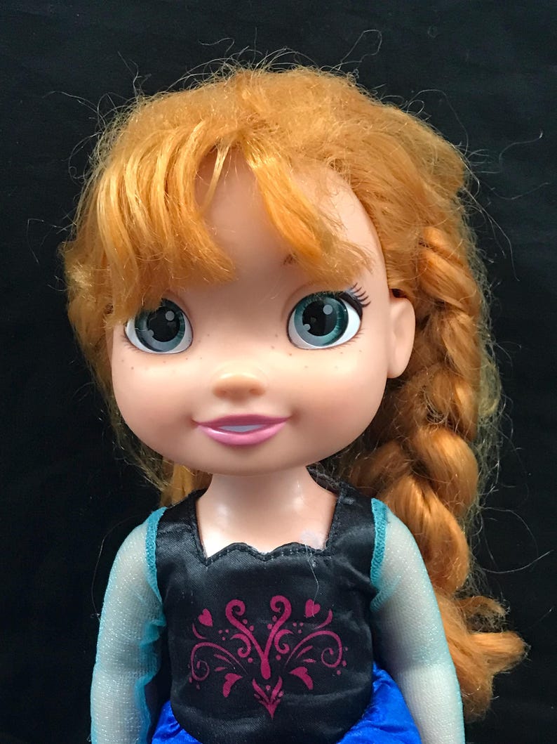 May include: A doll with orange hair styled in a side braid. The doll has blue eyes and is wearing a black top with a pink and purple design and a blue dress. The doll's skin is fair with freckles.