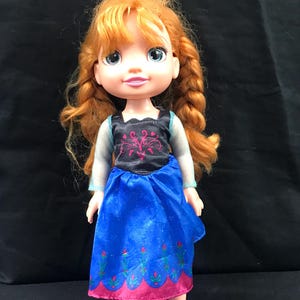 May include: A doll with red hair styled in braids, wearing a blue dress with a black bodice and pink accents. The doll has blue eyes and is wearing pink shoes. The doll is a character from the movie Frozen.