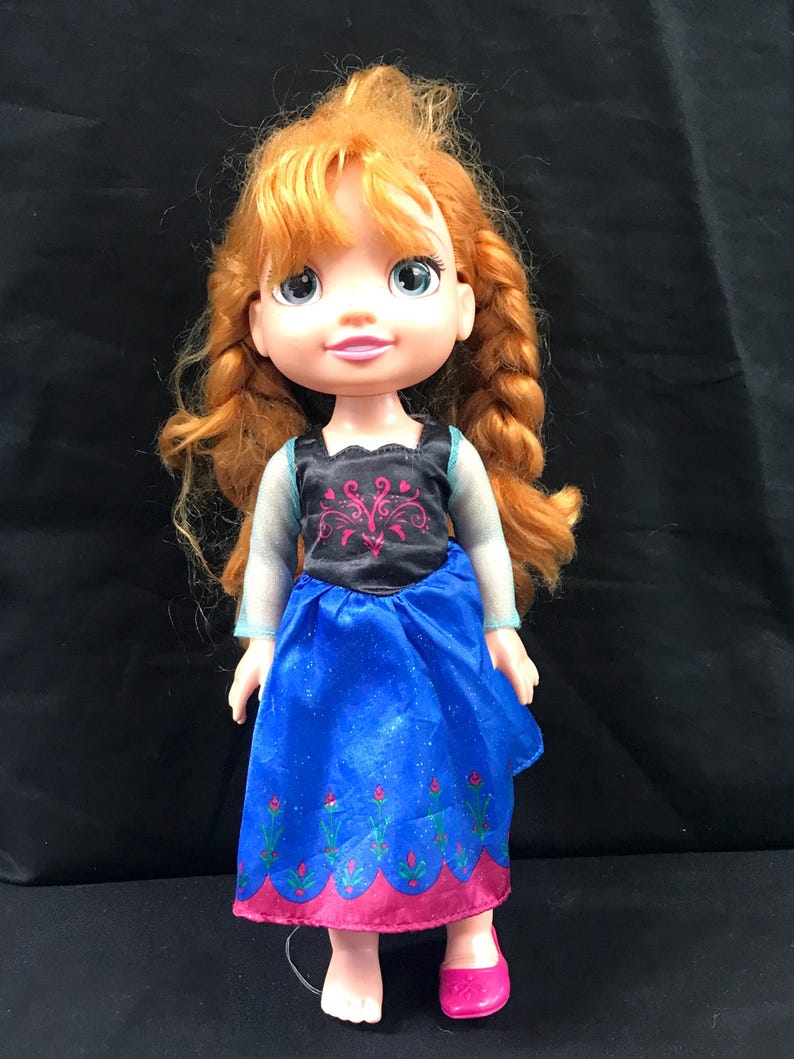 May include: A doll with red hair styled in braids, wearing a blue dress with a black bodice and pink accents. The doll has blue eyes and is wearing pink shoes. The doll is a character from the movie Frozen.