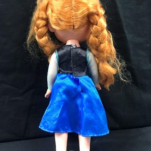 May include: A doll with orange braided hair, wearing a blue dress with a black bodice and pink shoes. The doll's back is visible, showcasing the details of the outfit and hairstyle. The doll is standing on a black surface.