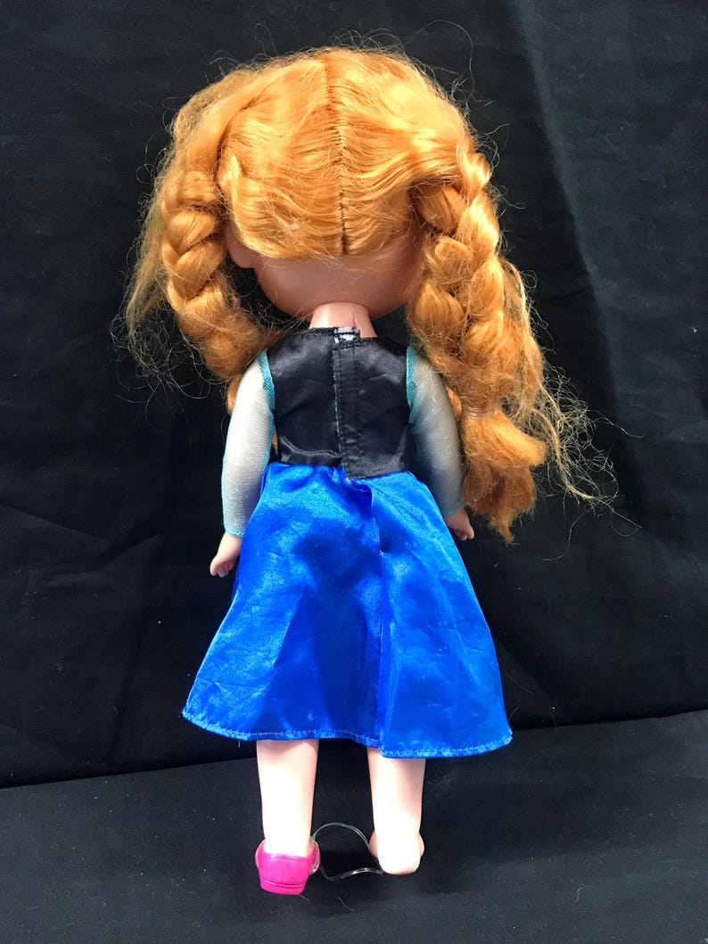 May include: A doll with orange braided hair, wearing a blue dress with a black bodice and pink shoes. The doll's back is visible, showcasing the details of the outfit and hairstyle. The doll is standing on a black surface.
