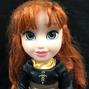 Puede incluir: Mu&ntilde;eca con cabello rojo y ojos azules, vestida con un vestido negro con detalles dorados. El vestido tiene un cintur&oacute;n marr&oacute;n y un cuello dorado. La mu&ntilde;eca tiene una expresi&oacute;n sonriente y pecas.