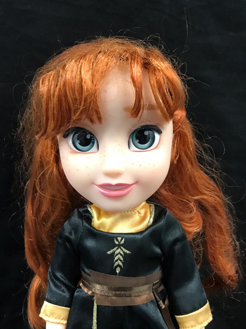 Puede incluir: Mu&ntilde;eca con cabello rojo y ojos azules, vestida con un vestido negro con detalles dorados. El vestido tiene un cintur&oacute;n marr&oacute;n y un cuello dorado. La mu&ntilde;eca tiene una expresi&oacute;n sonriente y pecas.