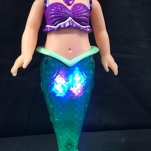 May include: A doll of a mermaid with red hair, wearing a purple top and a teal tail. The tail lights up with blue and purple lights. The doll has a white necklace with a purple seashell pendant.