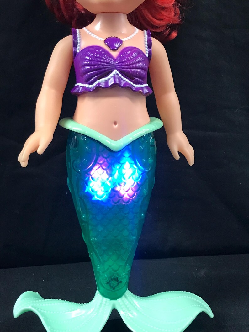 May include: A doll of a mermaid with red hair, wearing a purple top and a teal tail. The tail lights up with blue and purple lights. The doll has a white necklace with a purple seashell pendant.