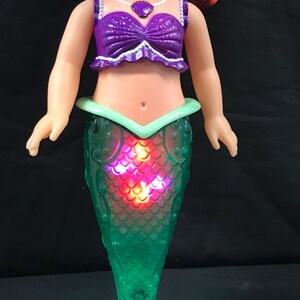 May include: A doll of a mermaid with red hair, wearing a purple top and a green tail. The tail lights up with red and orange lights. The doll has a light green tail fin and a purple seashell necklace.