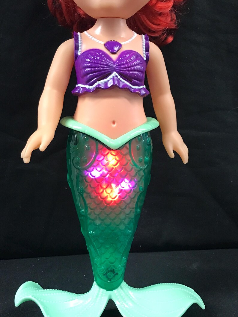 May include: A doll of a mermaid with red hair, wearing a purple top and a green tail. The tail lights up with red and orange lights. The doll has a light green tail fin and a purple seashell necklace.