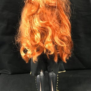 Puede incluir: Una mu&ntilde;eca con pelo largo y rizado de color naranja. La mu&ntilde;eca lleva un atuendo negro con botas negras. Las botas tienen detalles dorados. La mu&ntilde;eca se ve desde atr&aacute;s.