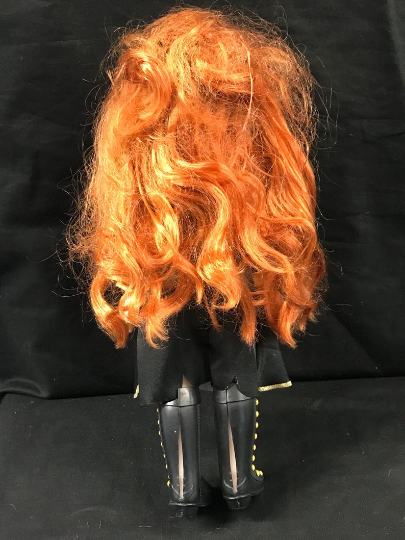 Puede incluir: Una mu&ntilde;eca con pelo largo y rizado de color naranja. La mu&ntilde;eca lleva un atuendo negro con botas negras. Las botas tienen detalles dorados. La mu&ntilde;eca se ve desde atr&aacute;s.