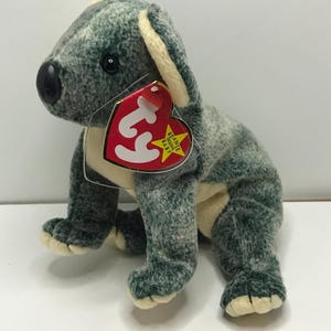 以下が含まれることがあります： 緑とクリーム色のぬいぐるみコアラ。 コアラは黒い鼻と目をしており、白い文字が入った赤いタグが付いています。 おもちゃは座った姿勢です。 子供のおもちゃ。