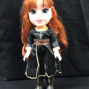 Puede incluir: Mu&ntilde;eca con cabello rojo y ojos azules, vestida con un vestido negro con ribetes dorados y un cintur&oacute;n marr&oacute;n. El vestido tiene dise&ntilde;os dorados en forma de hojas. Las botas de la mu&ntilde;eca son negras con detalles dorados.
