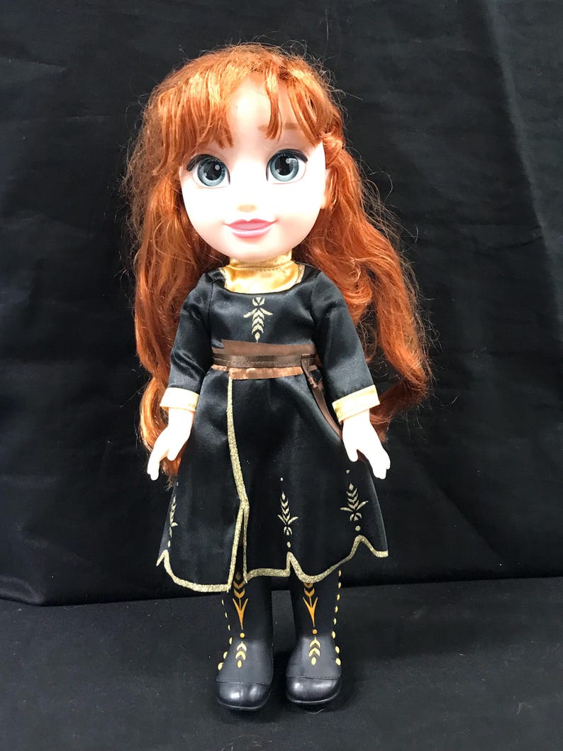 Puede incluir: Mu&ntilde;eca con cabello rojo y ojos azules, vestida con un vestido negro con ribetes dorados y un cintur&oacute;n marr&oacute;n. El vestido tiene dise&ntilde;os dorados en forma de hojas. Las botas de la mu&ntilde;eca son negras con detalles dorados.