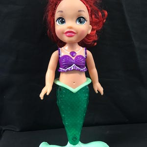 May include: A doll of a mermaid with red hair, blue eyes, and a purple top. The doll has a green mermaid tail and a light green fin. The doll is standing on a black surface.