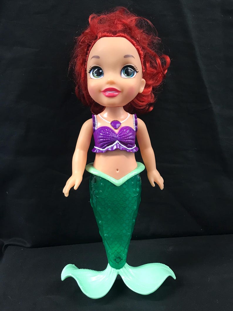 May include: A doll of a mermaid with red hair, blue eyes, and a purple top. The doll has a green mermaid tail and a light green fin. The doll is standing on a black surface.
