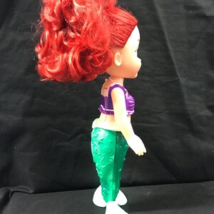 May include: A doll of a mermaid with bright red hair styled in a high ponytail. The doll wears a purple top and a green tail. The mermaid doll has white fins and is standing.