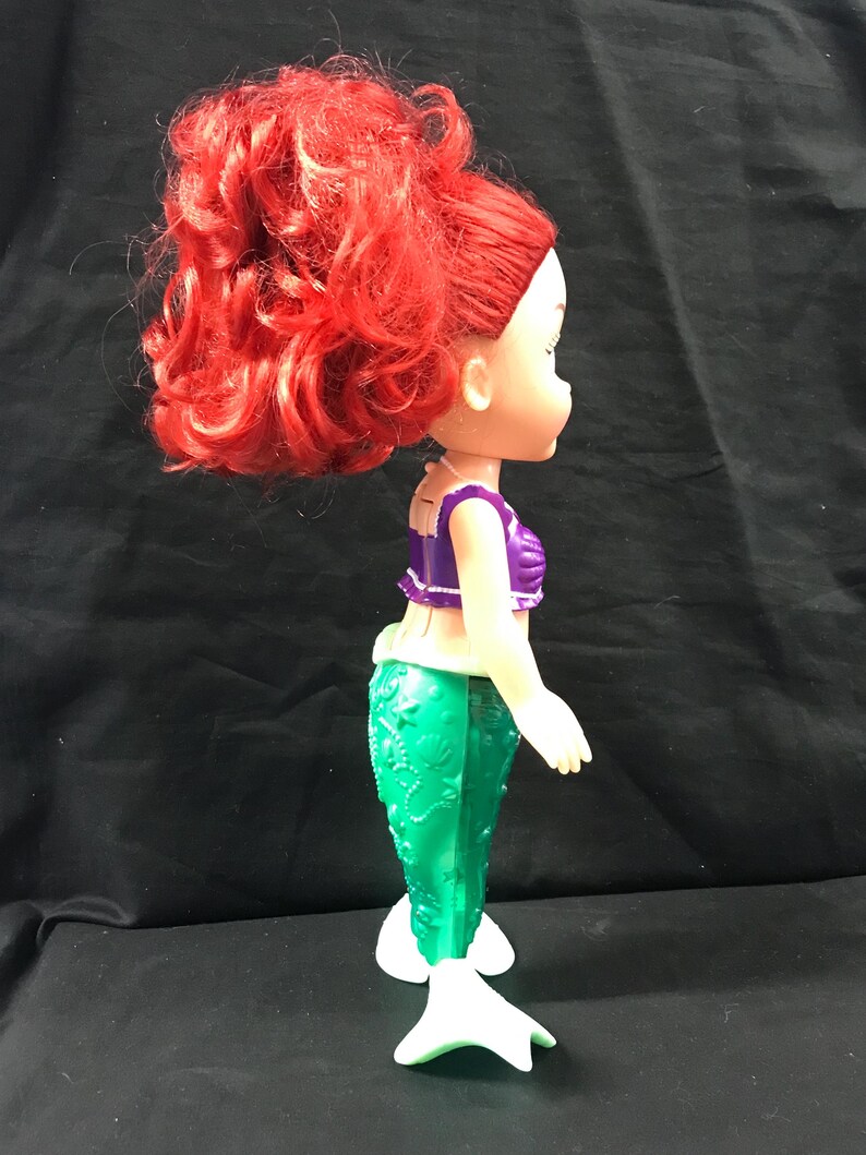 May include: A doll of a mermaid with bright red hair styled in a high ponytail. The doll wears a purple top and a green tail. The mermaid doll has white fins and is standing.