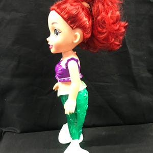 May include: A doll of a mermaid with red hair styled in a high ponytail. The doll wears a purple top, green tail, and white fins. The doll is standing and facing left.