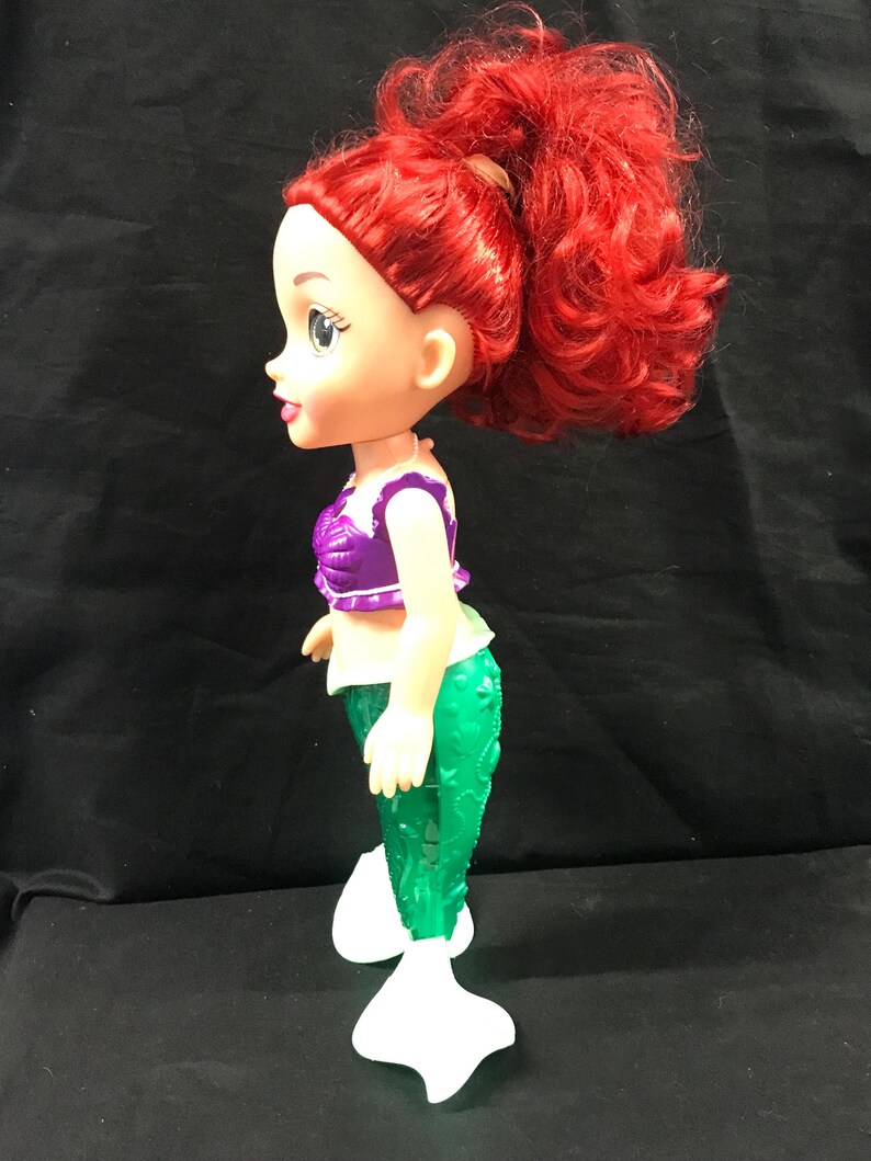 May include: A doll of a mermaid with red hair styled in a high ponytail. The doll wears a purple top, green tail, and white fins. The doll is standing and facing left.