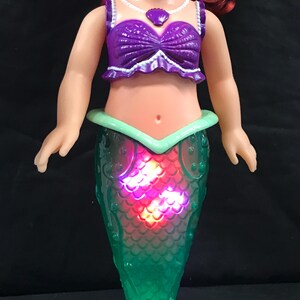 May include: A doll of a mermaid with red hair, wearing a purple top and a green tail. The tail lights up with red and pink lights. The doll has a white necklace with a purple pendant.