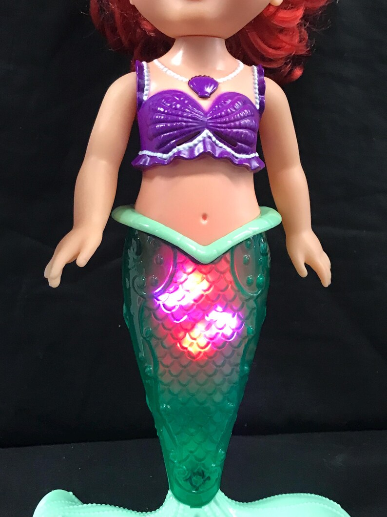 May include: A doll of a mermaid with red hair, wearing a purple top and a green tail. The tail lights up with red and pink lights. The doll has a white necklace with a purple pendant.