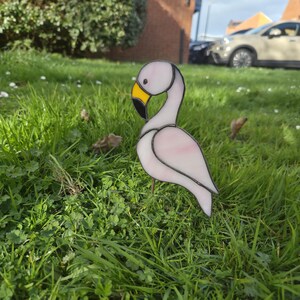 May include: A decorative stained glass flamingo garden ornament. The bird features a white body with pink detailing, a yellow beak with a black tip, and black outlines. It is set in a grassy garden.