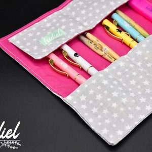 Puede incluir: Un estuche para lápices de tela rosa con un forro de tela gris con estrellas blancas. El estuche está abierto y tiene varios bolígrafos y lápices dentro.