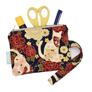 Puede incluir: Un estuche con cremallera con un diseño floral y de gato. El estuche es azul marino con flores rojas, flores color crema y una aplicación de gato blanco. Tijeras amarillas, bolígrafo azul y rotulador amarillo visibles.