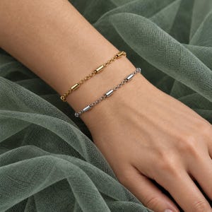 May include: Two delicate chain bracelets worn together on a wrist. One bracelet is gold-toned and the other is silver-toned. Both feature a series of small, smooth, cylindrical metal beads spaced evenly along the length of the fine link chains.