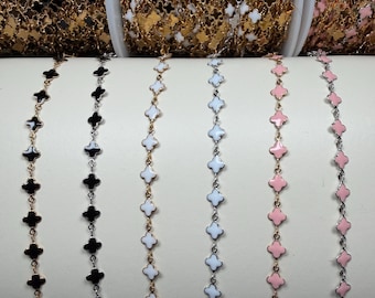 May include: Six metal chains featuring repeating clover-shaped charms in various colors including black, white, and pink. The chains are displayed on spools and draped over a curved support, showcasing gold and silver-toned metal links and enamel.