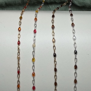 May include: Four vertical jewelry chains featuring small, oval-shaped beads in various colors including red, orange, yellow, brown, and clear. Two chains are gold-toned and two are silver-toned, each consisting of repeating metal links and beads.
