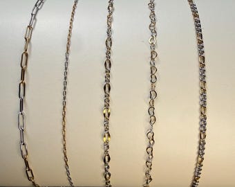 May include: Five distinct metal chains displayed vertically on a cylindrical white stand. The collection includes a gold and silver paperclip chain, a thin gold cable chain, a textured gold and silver link chain, a heart-link chain, and a curb chain.