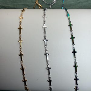 May include: Three delicate chains featuring small, evenly spaced cross charms. One chain is gold-toned, one is silver-toned, and one features an iridescent, multi-colored metallic finish. Each chain is draped vertically over a smooth, curved surface.