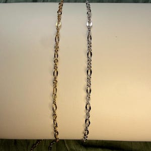 May include: Two delicate metal chains displayed vertically side by side. One chain is gold-toned and the other is silver-toned. Both chains feature a repeating pattern of elongated oval links alternating with smaller circular links throughout.
