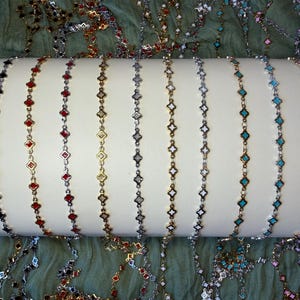 May include: A collection of eleven delicate metal chains draped over a white cylindrical display. Each chain features a repeating pattern of small, diamond-shaped enamel links in various colors including black, red, gold, silver, white, and blue.