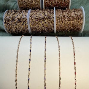 May include: Five spools of gold-toned metal chain, each featuring small, evenly spaced enamel accents in distinct colors including white, red, blue, purple, and pink. The chains are displayed on white spools and draped over a white cylindrical form.