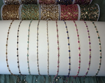 May include: A collection of gold-toned metal chains featuring small, evenly spaced enamel beads in various colors including black, light blue, white, pink, dark blue, magenta, and green, displayed on white spools and a white cylindrical stand.
