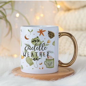May include: White ceramic mug with a gold handle. The mug has a white background with a pattern of green leaves, orange flowers, a pine cone, a star, a crescent moon, a teapot, a mitten, and a cup of hot chocolate. The text "cuddle weather" is printed in black on the mug.