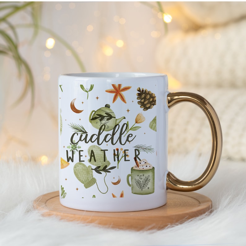 May include: White ceramic mug with a gold handle. The mug has a white background with a pattern of green leaves, orange flowers, a pine cone, a star, a crescent moon, a teapot, a mitten, and a cup of hot chocolate. The text "cuddle weather" is printed in black on the mug.