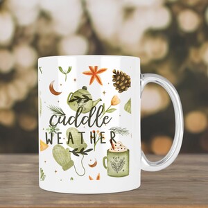 May include: White ceramic mug with a handle. The mug has a design of green leaves, a teapot, a pine cone, a star, a crescent moon, and a cup of hot chocolate. The text "cuddle weather" is written in black on the mug.