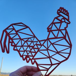 May include: A red geometric rooster cutout made of wood. The rooster is in a geometric style with a detailed design. The rooster is held in a hand against a blue sky and green grass background.