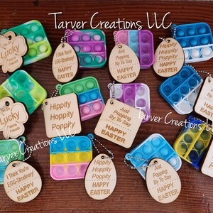 May include: A collection of 10 Easter-themed pop-it fidget toys with wooden tags. The tags have various Easter-themed messages, including "Just Popping By To Say Happy Easter", "Hippity Hoppity Poppity", and "I Think You're EGG-Strodinary! Happy Easter".