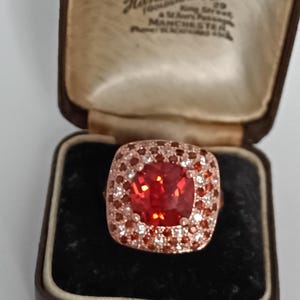 Op de afbeelding: Een roségouden ring met een grote, vierkant geslepen rode edelsteen, omringd door kleinere stenen. De ring wordt gepresenteerd in een vintage sieradendoosje met de tekst "Hancock & Son".