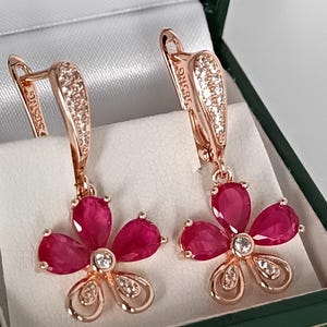 Könnte beinhalten: Ein Paar roségoldene Blumenohrringe mit rubinroten Blütenblättern und kleinen, klaren Akzentsteinen. Die Ohrringe werden in einer grünen Schmuckschatulle mit weißem Innenraum präsentiert. Blumen-Design.