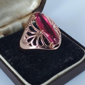 Op de afbeelding: Roségouden ring met een grote, rechthoekige, robijnkleurige edelsteen. De ring heeft een open ontwerp met een bloemmotief. De ring wordt gepresenteerd in een vintage sieradendoosje met een donker interieur.