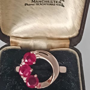 Op de afbeelding: Een roségouden ring met drie ronde, felrode edelstenen in een halvemaanvorm, geaccentueerd door kleine heldere steentjes. De ring wordt gepresenteerd in een donkerbruin, scharnierend juwelendoosje met een crèmekleurige voering en tekst.