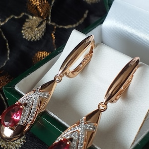 Könnte beinhalten: Ein Paar Ohrringe aus Roségold mit tropfenförmigen roten Edelsteinen und einem diamantartigen Design. Die Ohrringe werden in einer grün-weißen Schachtel präsentiert.