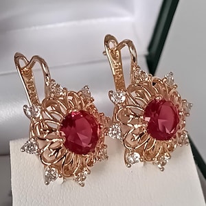 May include: Rose gold earrings featuring a central, round, red gemstone surrounded by a floral filigree design. Small, clear gemstones are set around the perimeter. The earrings have a lever back closure.
