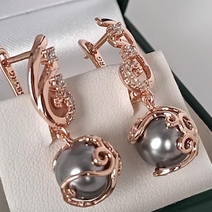 May include: Rose gold earrings with grey pearl drops. The earrings have a delicate design with small clear stones and a swirling pattern around the pearls. The earrings are displayed in a green and white box.
