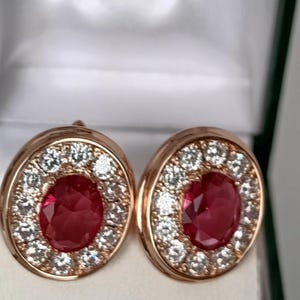 Puede incluir: Un par de pendientes de botón de forma ovalada engastados en oro rosa. Cada pendiente presenta una piedra preciosa de color rojo intenso con talla ovalada en el centro, rodeada por un halo de pequeñas piedras blancas redondas y claras.